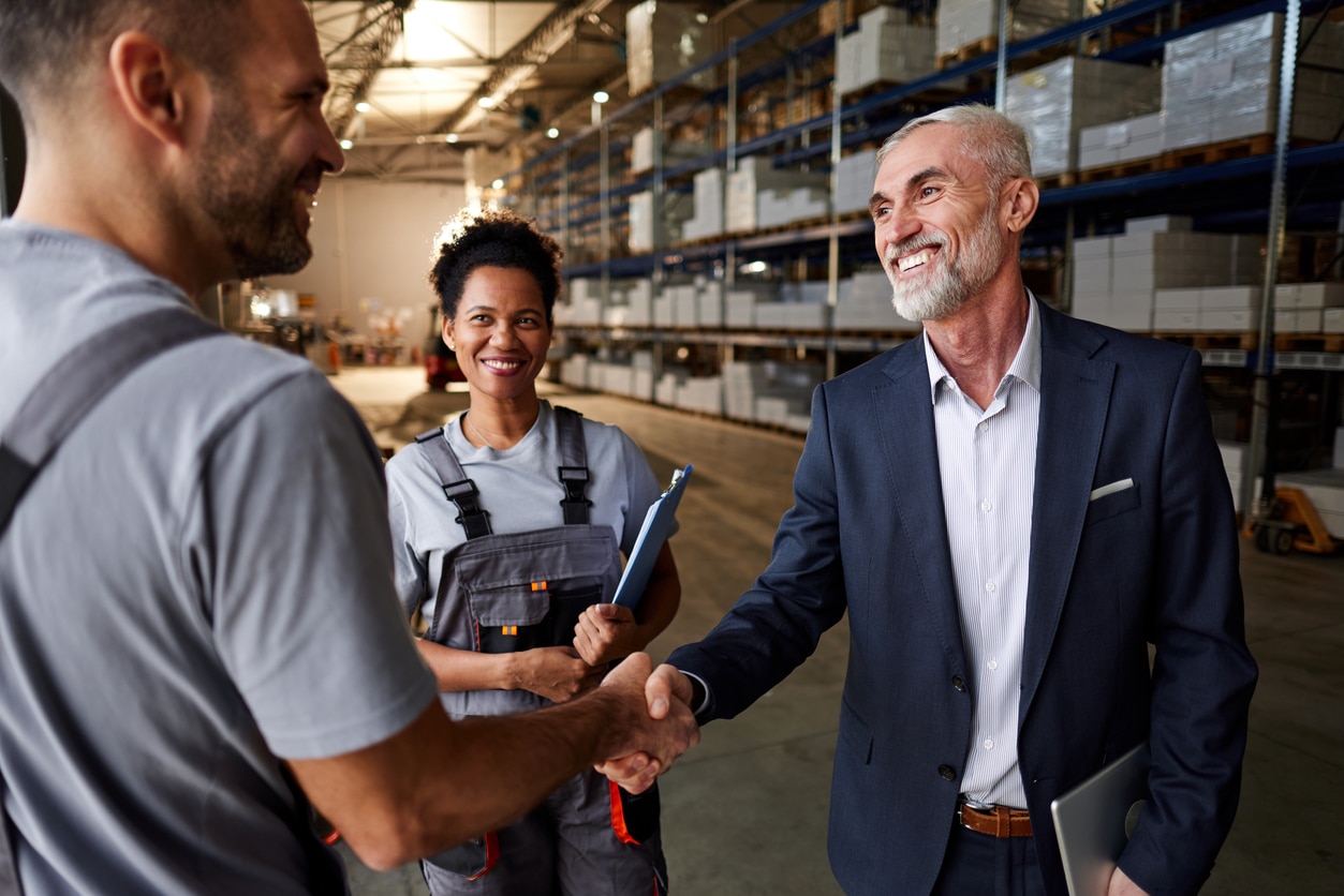 Happy manager and manual worker shaking hands next to black female colleague in distribution warehouse.